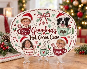May include: An oval ceramic platter featuring the text Grandma's Hot Cocoa Crew, decorated with illustrations of candy canes, snowflakes, and four mugs containing photos of children and a dog, each wearing a Santa hat.