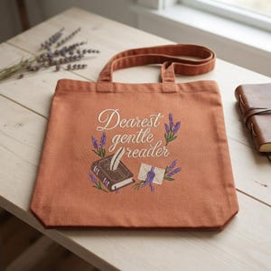 May include: A rust-coloured canvas tote bag featuring an embroidered design of a brown book with a white quill, a sealed envelope, and purple lavender sprigs. The text "Dearest gentle reader" is embroidered in white script above the items.