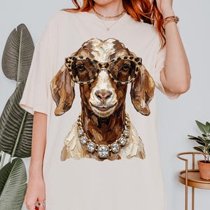 May include: A cream-colored t-shirt featuring a graphic illustration of a brown goat wearing leopard-print glasses and a chunky rhinestone necklace. The goat is depicted with textured, painterly brushstrokes in shades of brown and white.