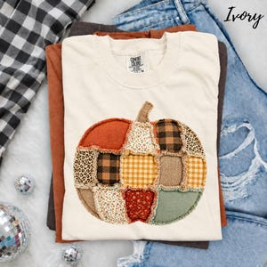May include: An ivory short-sleeve t-shirt featuring a large pumpkin graphic made of various fabric patches. The pumpkin design includes patterns such as leopard print, black and white buffalo check, yellow gingham, and floral prints in red and white.