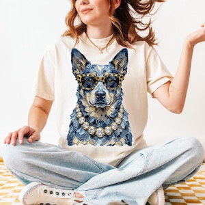 May include: A cream-colored t-shirt featuring a graphic of a blue cattle dog wearing leopard-print glasses and a chunky diamond-style necklace. The dog illustration is rendered in a textured, painterly style with visible brushstrokes in shades of blue.