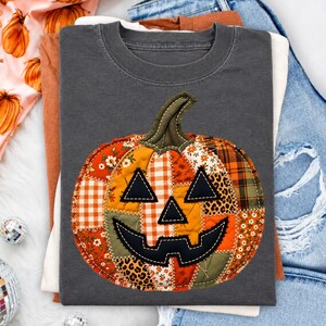 May include: A charcoal gray t-shirt featuring a large, central pumpkin graphic. The pumpkin is constructed from various fabric patches in orange, white, and green, displaying patterns like gingham, floral, and leopard print with black stitched eyes.