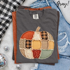 May include: A dark gray crew neck t-shirt featuring a large pumpkin graphic made of various fabric patches including leopard print, black and white buffalo check, yellow gingham, and floral patterns with frayed edges. The shirt has a Comfort Colors label.
