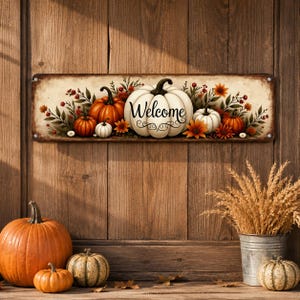 May include: A rectangular metal sign featuring a cream-colored background with a large central white pumpkin, smaller orange and white pumpkins, and autumn foliage. The word Welcome is printed in black script across the center of the large pumpkin.