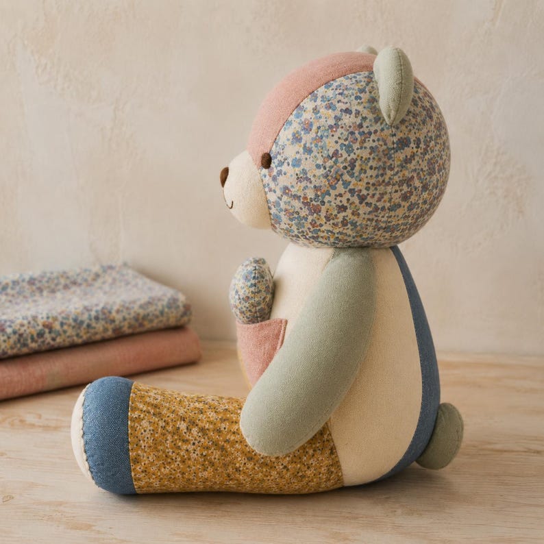 Op de afbeelding: Een knuffelbeer gemaakt van diverse stoffen lappen, waaronder bloemmotieven in blauw, geel en roze, naast effen delen in cr&egrave;me, saliegroen en spijkerstofblauw. De beer heeft een klein zakje op de borst.