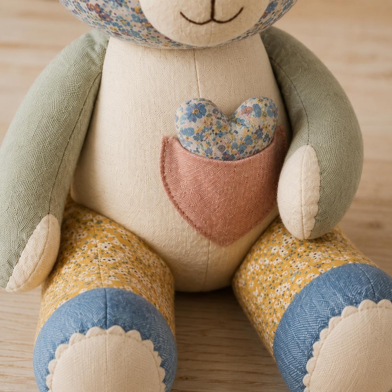 Op de afbeelding: Een knuffelbeer met een cr&egrave;mekleurige romp, een roze zakje met een blauw bloemenhart erin. De armen zijn saliegroen, de benen hebben een geel bloemenpatroon met blauwe boorden en het hoofd heeft oren met een blauw bloemenpatroon.