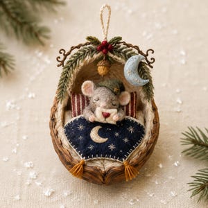 May include: A felted mouse ornament nestled inside a walnut shell, wearing a green cap and holding a small envelope. The mouse is tucked under a dark blue blanket with a crescent moon and stars, resting against red and white striped pillows.
