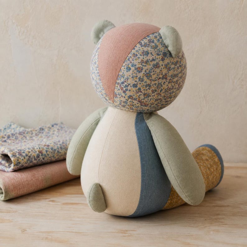 Op de afbeelding: Een knuffelbeer gemaakt van patchwork stoffen panelen in diverse kleuren en patronen, waaronder een bloemenprint, effen saliegroen, spijkerblauw, cr&egrave;me en mosterdgeel, voorzien van een klein staartje en ronde oren.