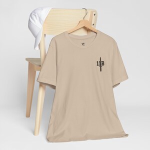 May include: A beige short-sleeved t-shirt featuring a small black graphic on the left chest area with the text 11B and a dagger icon. A plain white baseball cap is draped over the top of the wooden chair back behind the t-shirt.