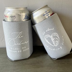 May include: Two gray can coolers with white text. One says "The Denhunt" and "Biloxi, MS February 22, 2025." The other says "it's about shucking time" with an oyster graphic. Each cooler holds a silver can.