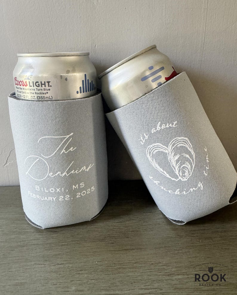May include: Two gray can coolers with white text. One says "The Denhunt" and "Biloxi, MS February 22, 2025." The other says "it's about shucking time" with an oyster graphic. Each cooler holds a silver can.