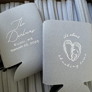 May include: Gray drink koozies with white text. One koozie reads "The Derkins, Biloxi, MS, February 22, 2025." The other says "it's about shucking time" with a heart-shaped oyster design. The koozies are stacked in a box.