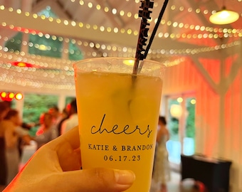 May include: A clear plastic cup with a yellow drink and two straws. The cup has the text "cheers" printed on it, along with the names "Katie & Brandon" and the date "06.17.23".