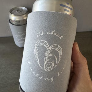 May include: A gray can cooler with the text "its about fucking time" and a white oyster shell design. A silver can is inside the cooler. Another can cooler with a silver can is in the background.