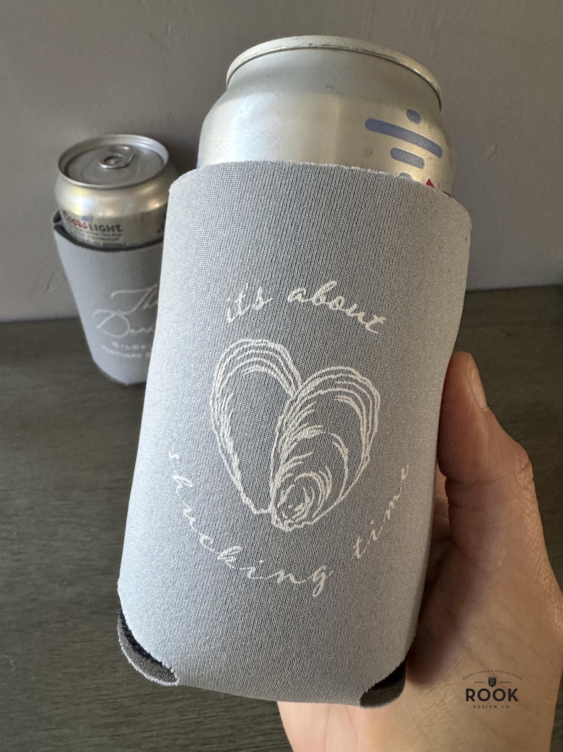 May include: A gray can cooler with the text "its about fucking time" and a white oyster shell design. A silver can is inside the cooler. Another can cooler with a silver can is in the background.