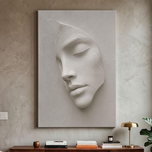 Op de afbeelding: Een rechthoekig wandkunstwerk met een monochroom lichtgrijs reliëfsculptuur van een menselijk gezicht met gesloten ogen. Het oppervlak heeft een textuur die lijkt op penseelstreken, wat diepte en schaduw creëert over de gelaatstrekken.