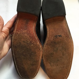 Puede incluir: Primer plano de las suelas de cuero marr&oacute;n de un par de zapatos de cuero negro. Las suelas tienen un borde cosido y el texto "Made in England" es visible en el tal&oacute;n.
