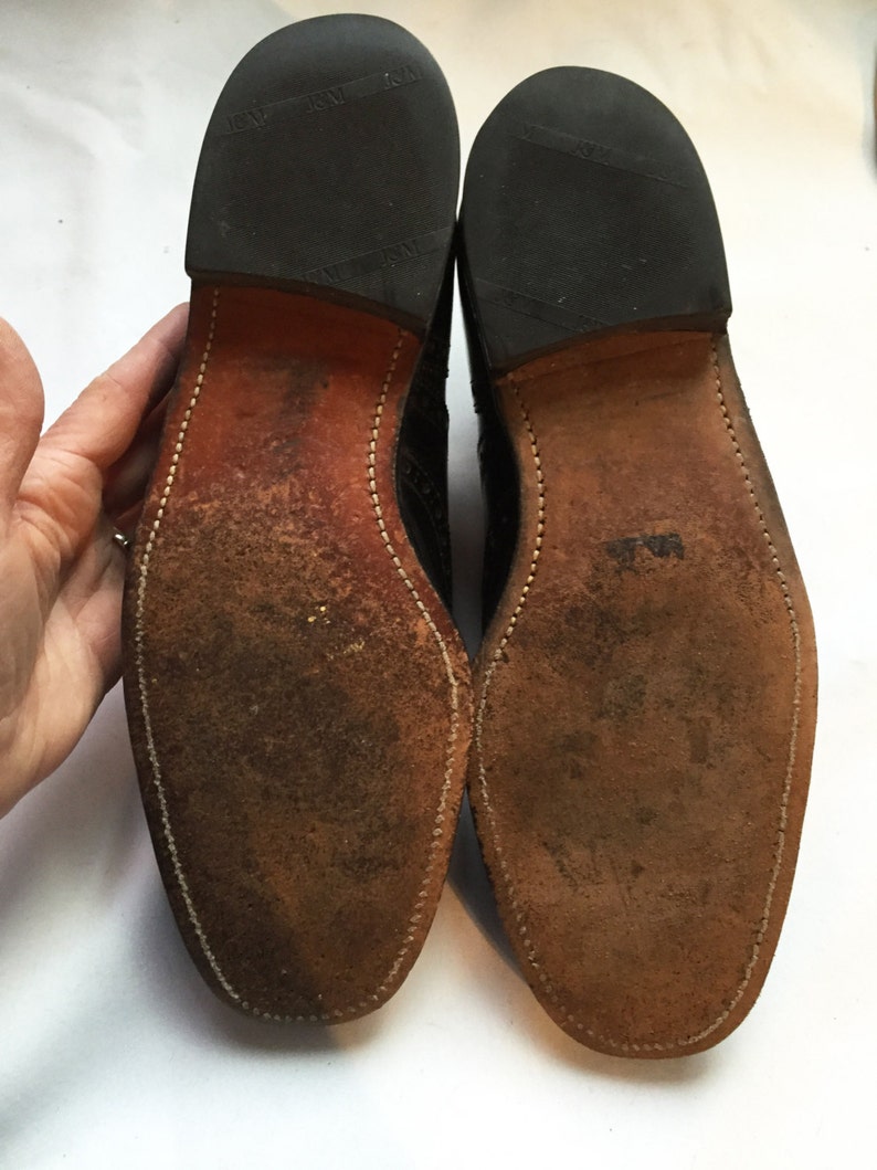 Puede incluir: Primer plano de las suelas de cuero marr&oacute;n de un par de zapatos de cuero negro. Las suelas tienen un borde cosido y el texto "Made in England" es visible en el tal&oacute;n.
