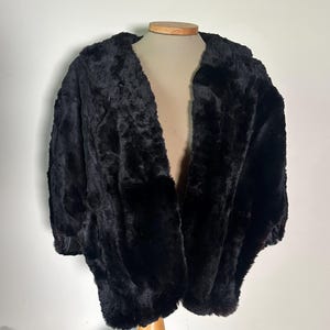 以下が含まれることがあります： 木製のマネキンに展示された黒いフェイクファーコート。 コートは幅広のオープンフロントと長袖です。 毛皮は厚く、ふわふわに見え、わずかにテクスチャード加工されています。 コートは無地の黒色です。