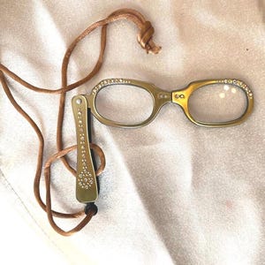 May include: Gold-toned reading glasses with a decorative handle and a brown cord. The frames are embellished with small, sparkling rhinestones. The glasses are designed to be worn around the neck.