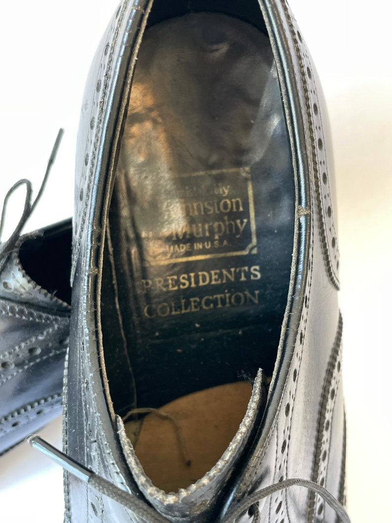 Puede incluir: Zapatos de vestir de cuero negro con dise&ntilde;o de punta de ala y cierre de cordones. Los zapatos tienen una etiqueta que dice "Johnston & Murphy Presidents Collection Made in U.S.A."