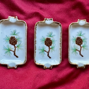 Pode incluir: Três cinzeiros retangulares brancos com acabamento dourado e um desenho de pinha. Cada cinzeiro tem um pequeno entalhe numa extremidade. A pinha é castanha, com agulhas verdes e um caule castanho.