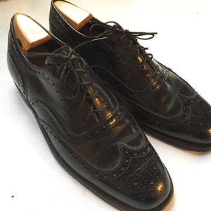 Puede incluir: Un par de zapatos de vestir negros de cuero con cordones. Los zapatos tienen un dise&ntilde;o de punta de ala y est&aacute;n hechos de cuero liso y pulido.