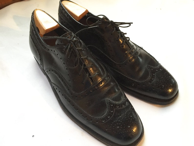 Puede incluir: Un par de zapatos de vestir negros de cuero con cordones. Los zapatos tienen un dise&ntilde;o de punta de ala y est&aacute;n hechos de cuero liso y pulido.