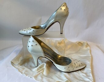 May include: A pair of white satin high heels with a pointed toe and a rhinestone embellishment. The heels are on a white satin surface.