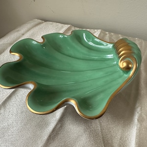 May include: A decorative, seafoam green ceramic shell-shaped dish with gold trim. The dish has a scalloped edge and a textured, ribbed interior. The gold accents highlight the edges and the curled end of the shell, creating an elegant accent piece.