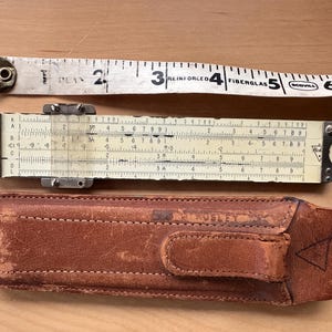 May include: An overhead shot of a vintage collection of measuring tools. A white and black reinforced fiberglass tape measure is above a slide rule and a brown leather case. The slide rule is cream-colored with black markings.