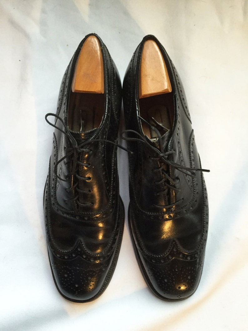 Puede incluir: Un par de zapatos negros de cuero con cordones, punta de ala y patr&oacute;n de brogue.