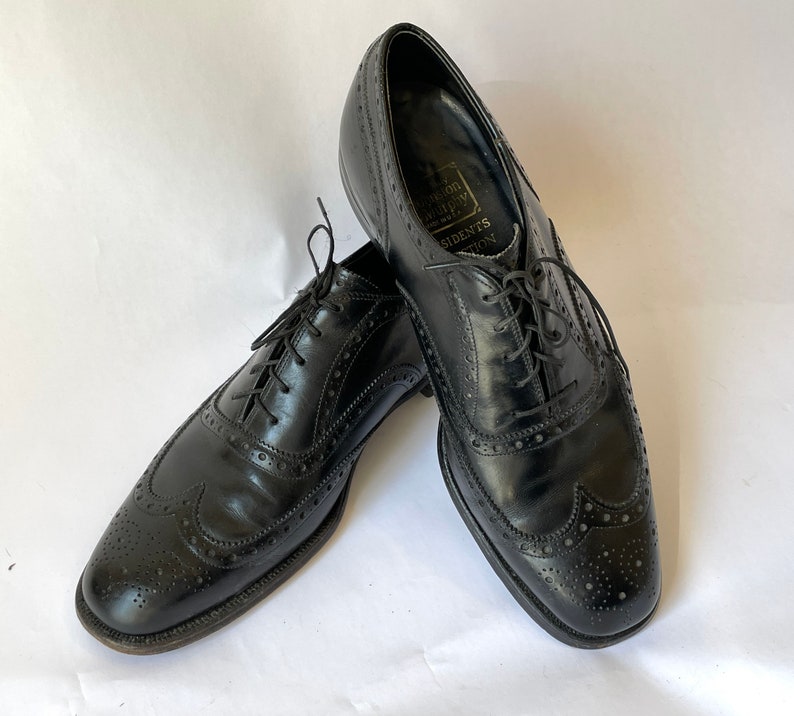 Puede incluir: Un par de zapatos negros de cuero con cordones y detalles de punta de ala. Los zapatos tienen un patr&oacute;n de brogue y est&aacute;n hechos por Johnston & Murphy.