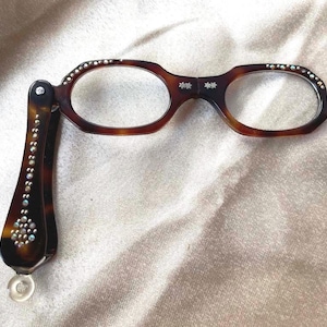 May include: Brown tortoiseshell reading glasses with a folding design. The frames are embellished with small, sparkling rhinestones along the top and on the arms. The bridge features two small floral designs.