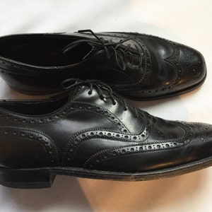 Puede incluir: Un par de zapatos Oxford de cuero negro con detalles de punta de ala. Los zapatos tienen cierre de cordones y un patr&oacute;n de brogue.