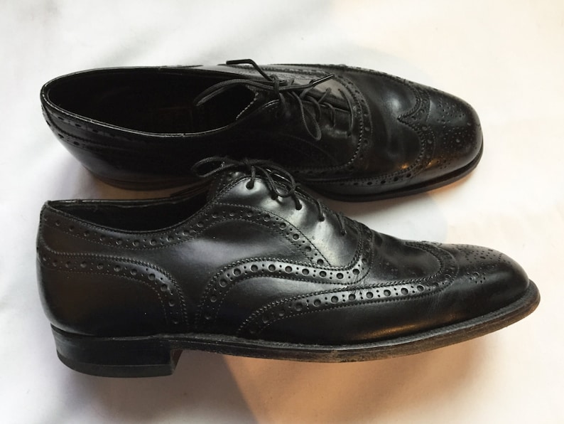 Puede incluir: Un par de zapatos Oxford de cuero negro con detalles de punta de ala. Los zapatos tienen cierre de cordones y un patr&oacute;n de brogue.