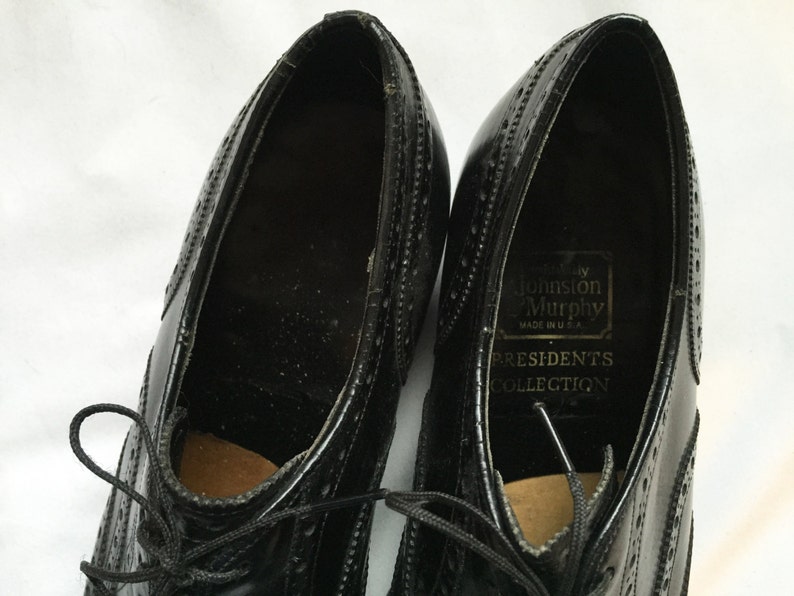 Puede incluir: Un par de zapatos negros de cuero con cordones y detalles de punta de ala. Los zapatos est&aacute;n etiquetados con "Johnson & Murphy" y "Presidents Collection" en el interior.