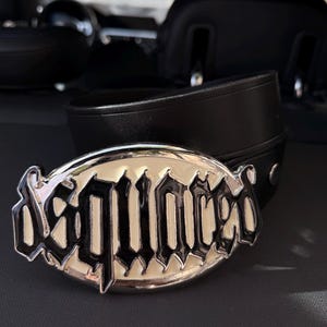 Peut inclure: Une ceinture en cuir noir dotée d'une grande boucle en métal argenté de forme ovale. La boucle affiche le mot Dsquared dans une police noire stylisée d'inspiration gothique sur un fond en émail crème avec un bord argenté poli.