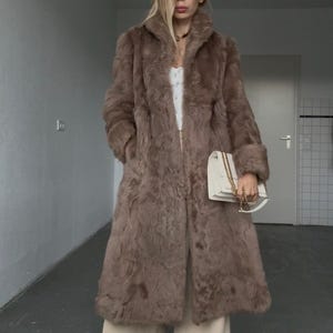 Può includere: Un lungo cappotto in pelliccia sintetica marrone con ampio colletto e maniche lunghe, indossato sopra un top bianco e pantaloni beige a gamba larga. Una borsa a tracolla rettangolare bianca con catena dorata è tenuta in mano, sneakers bianche.