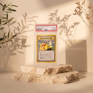 May include: A graded 1998 Japanese promo Pikachu Illustrator trading card in a clear plastic slab. The card features a yellow character holding a paintbrush, the text ILLUSTRATOR, and Japanese script. The label at the top reads 1998 P.M. JAPANESE PROMO PIKACHU-HOLO GEM MT.