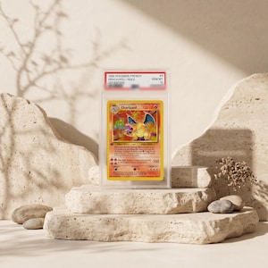 May include: A 1999 French 1st Edition Charizard trading card encased in a clear PSA protective slab. The card features a holographic illustration of the orange dragon-like creature, with text including Charizard, 120 HP, and fire energy symbols.