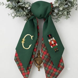 May include: A decorative fabric bow featuring a solid dark green front with an embroidered gold letter C on one side and a nutcracker figure on the other. The reverse side and edges display a red, green, and gold plaid pattern throughout.