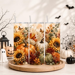 Puede incluir: Tres vasos cilíndricos con pajitas que presentan un diseño de temática otoñal. El estampado muestra calabazas naranjas y blancas, girasoles amarillos, hojas de otoño, bayas y mariposas sobre un fondo claro de estilo acuarela.