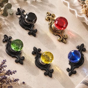 Può includere: Cinque pomelli decorativi per cassetti con pomelli in vetro sfaccettato dai toni gioiello in nero, rosso, verde, giallo e blu. Ogni pomello è montato su una piastra metallica decorata a volute, con finitura nero opaco o ottone antico.