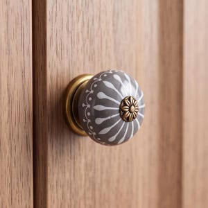 Puede incluir: Un pomo de armario redondo de cerámica con superficie gris, un patrón radial de pétalos blancos y un detalle floral central en tono latón. El pomo está montado sobre una base circular de tono latón, fijada a un panel de madera de color marrón claro.