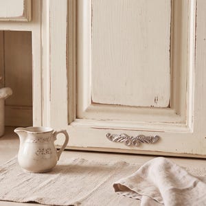 May include: A cream-colored ceramic creamer with a small handle and a subtle floral pattern sits on a textured beige fabric mat. Behind it is a cabinet door with a distressed off-white finish and an ornate, silver-toned metal drawer pull.