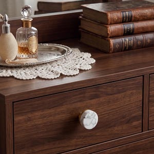 May include: A dark wood dresser featuring a round drawer knob with a white and grey marble pattern. On the surface sits a silver tray, a glass perfume bottle with a gold neck, a cream-colored textured atomizer, and a stack of three leather-bound books.