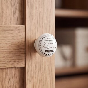 May include: A round white ceramic cabinet knob featuring a black circular border with small stars. The center displays the French text "Je pense donc je suis" in a cursive script, mounted on a light brown wood cabinet door with vertical grain.