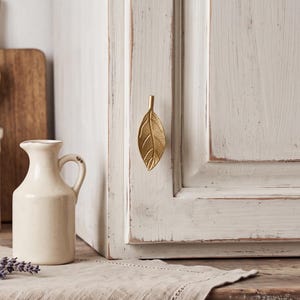 May include: A gold-toned metal cabinet knob shaped like a textured leaf, mounted on a white painted wooden cabinet door with a distressed finish. The leaf design features a central vein and fine detailing across the surface of the metal.