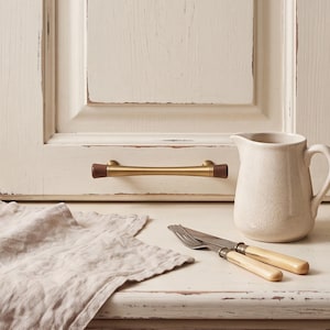 May include: A cabinet pull featuring a central brushed gold metal bar with dark brown wooden end caps, mounted horizontally on a distressed off-white cabinet door, accompanied by a ceramic pitcher and a fork and knife set with cream handles.
